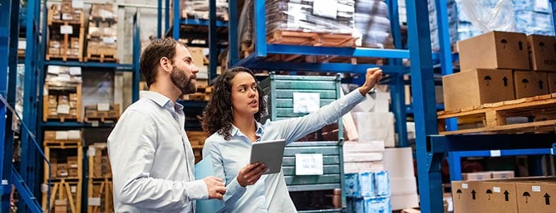 Mujer de negocios con una tableta en la mano. Habla con un hombre y le señala unos paquetes (el stock). Ambos están en un centro de distribución o warehouse